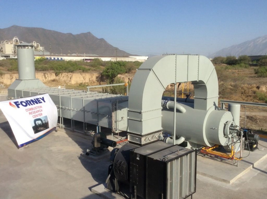 An outdoor view of the "FORNEY Combustion Research Center" test facility, featuring large grey industrial ducts, a round combustion chamber, and a mountain backdrop.