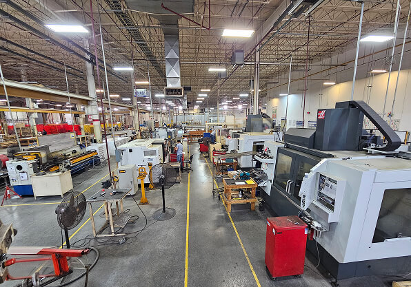 A wide interior shot of a well-lit, industrial machine shop featuring rows of CNC machinery and equipment stretching down a central aisle marked with yellow safety lines.